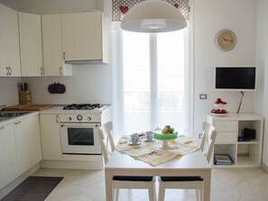 Una cocina con armarios blancos, estufa, horno, una mesa de comedor con sillas, una ventana con cortinas y un televisor montado en la pared.