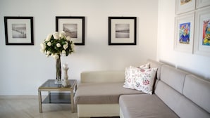 Ein Wohnzimmer mit einem beigen Sofa, einem Glastisch und einer Vase mit weißen Blumen. An der Wand hängen drei gerahmte Bilder.