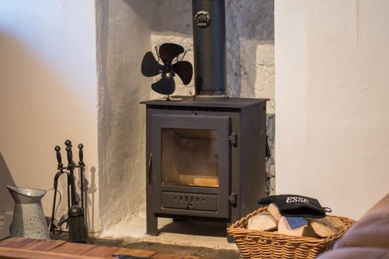 A wood-burning stove with a fan, a metal container, and a basket of firewood.
