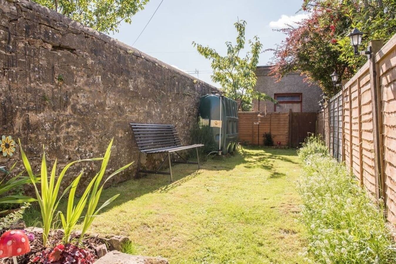 Un jardin avec un mur en pierre, un banc, un abri de jardin vert et une clôture en bois.