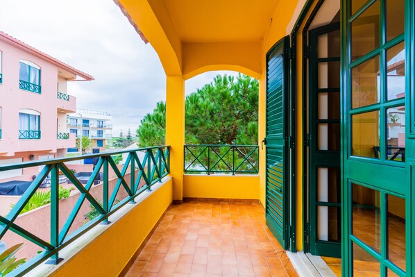 Un balcón con pared amarilla, barandilla verde y vista de edificios y árboles cercanos.