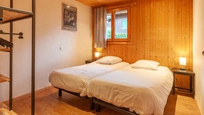 A bedroom with two beds, wooden walls, a window with curtains, and a small table with a lamp. - Top location in the resort center with easy access to slopes (Morzine)