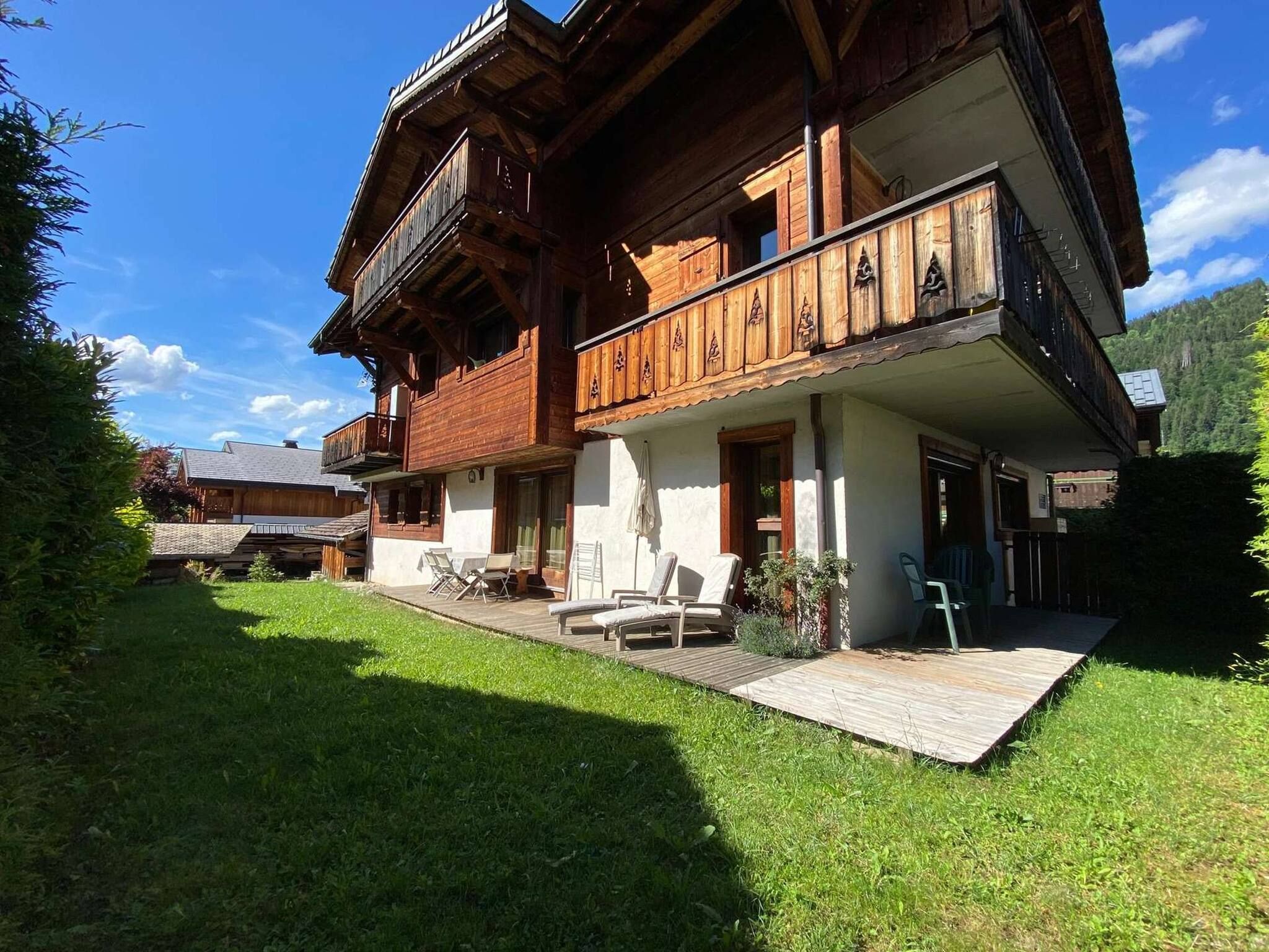 A wooden chalet with a balcony, a covered patio with lounge chairs, and a well-maintained lawn.