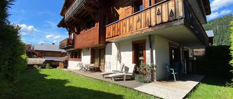 A wooden chalet with a balcony, a covered patio with lounge chairs, and a well-maintained lawn.