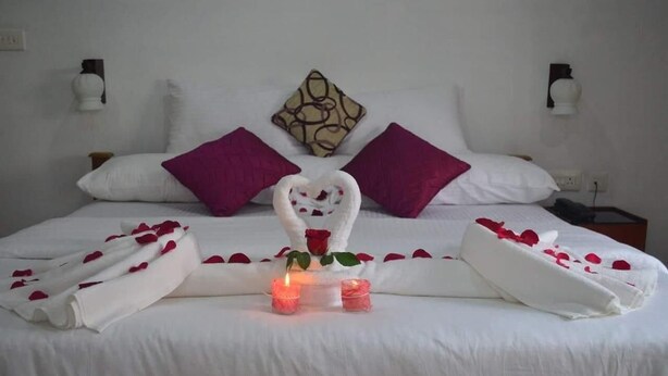 A neatly made bed with white linens, purple pillows, and a heart-shaped towel centerpiece. - Green Island Resort (Vaikom)