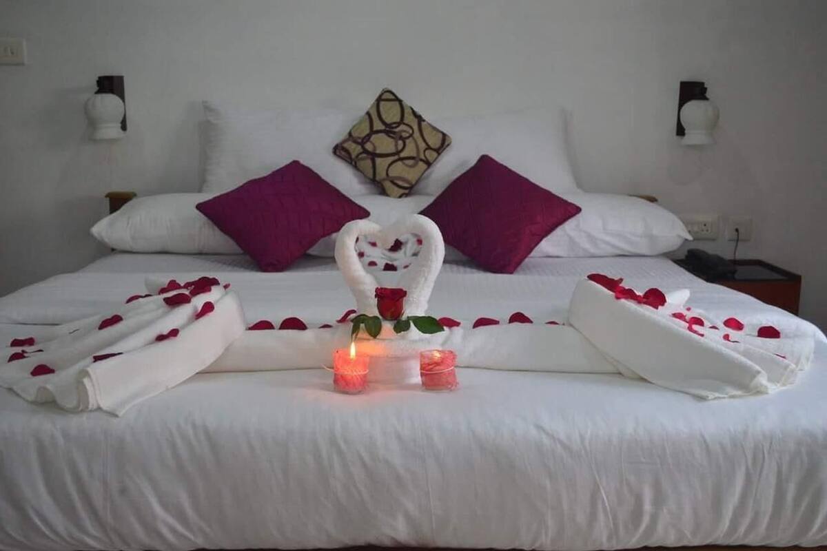 A neatly made bed with white linens, purple pillows, and a heart-shaped towel centerpiece.