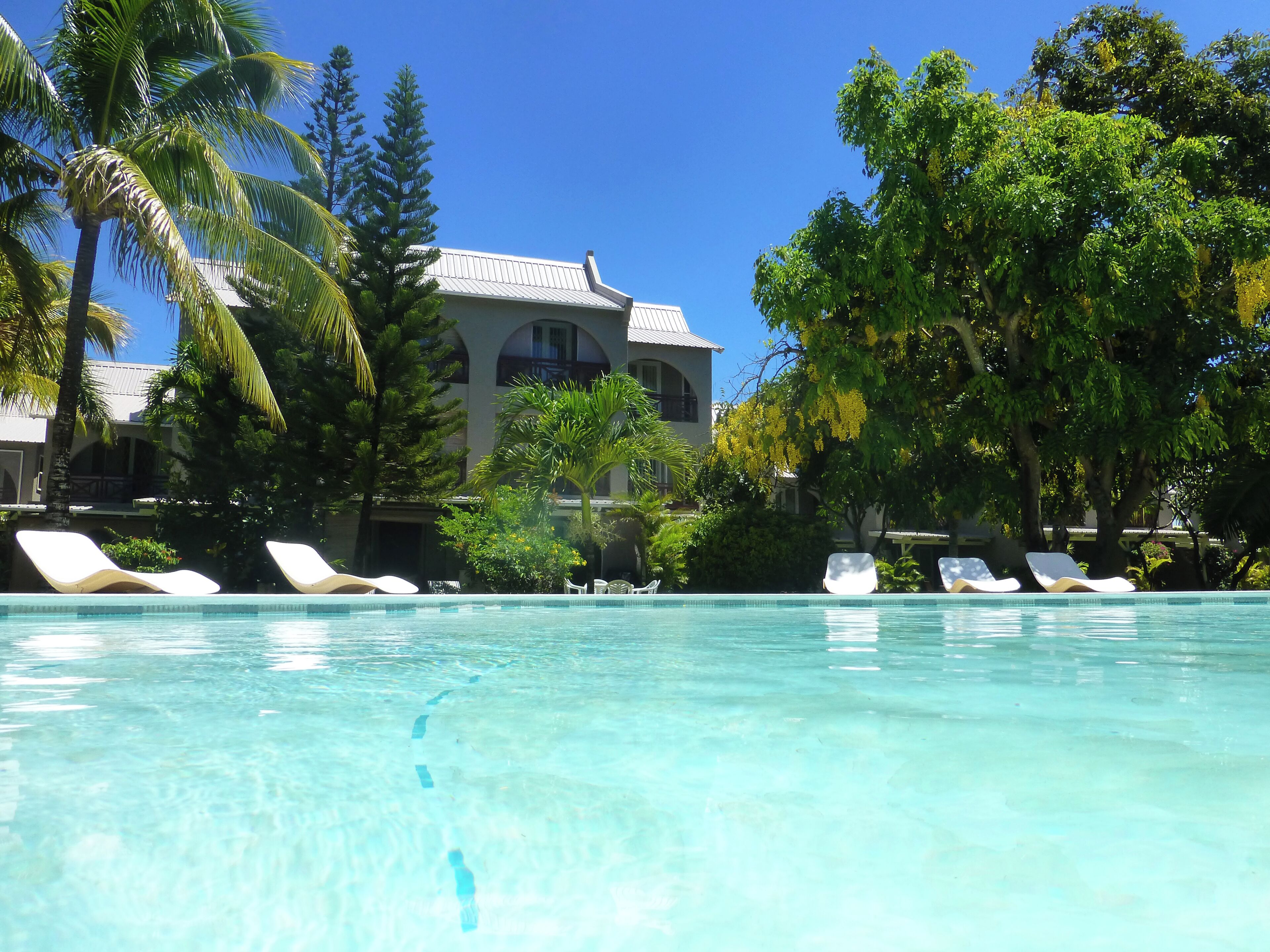 A clear pool with lounge chairs, surrounded by palm trees and a building with a tiled roof.