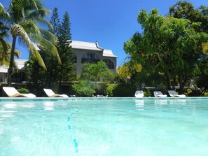 A clear pool with lounge chairs, surrounded by palm trees and a building with a tiled roof.