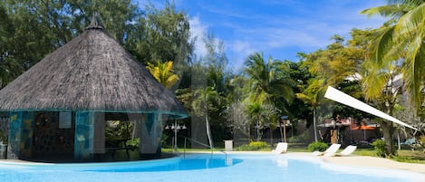 Una piscina con sillas de descanso, una cabaña con techo de paja y vegetación tropical.