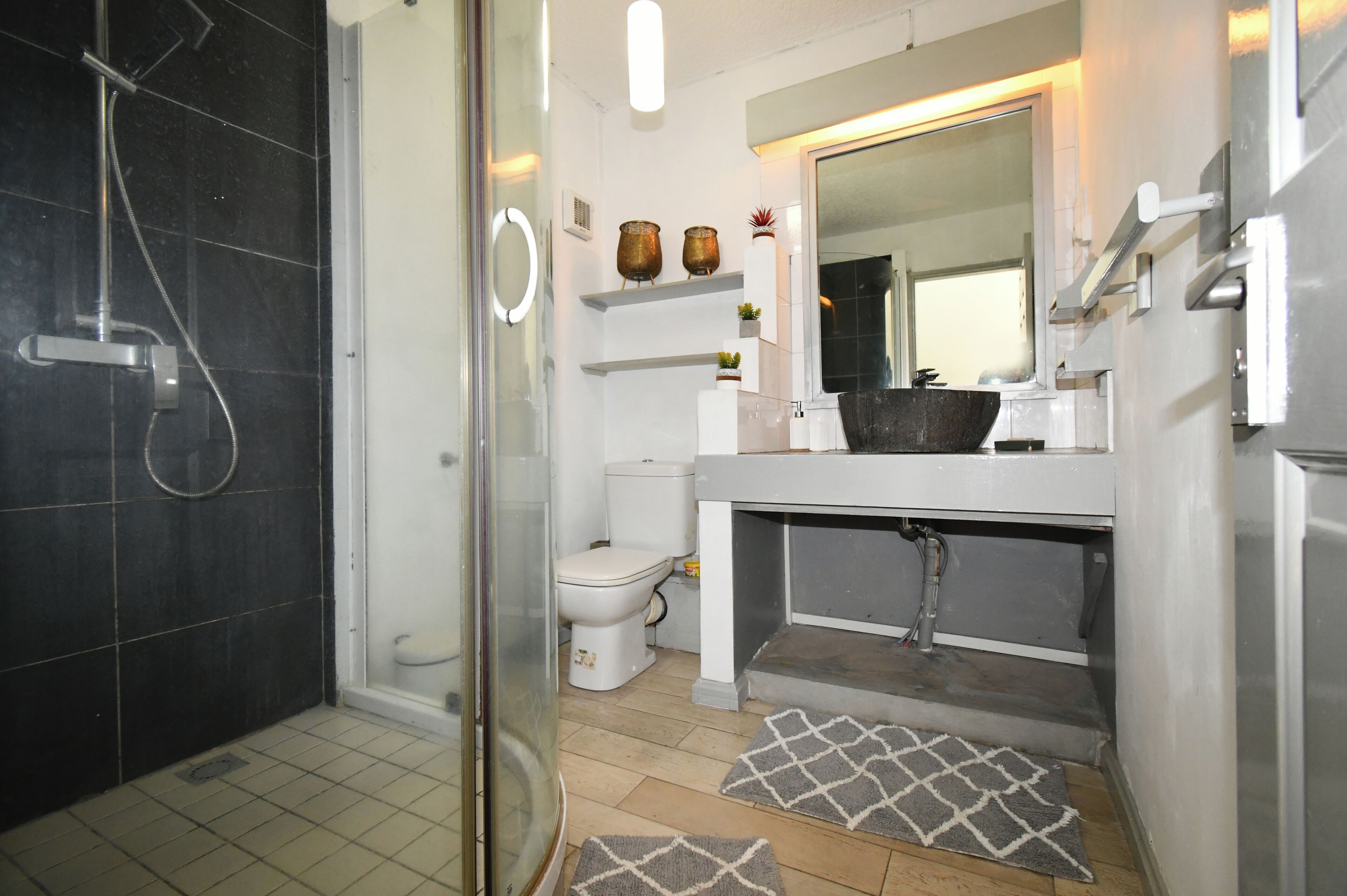 A bathroom with a glass shower enclosure, a toilet, a sink with a mirror, and a small shelf with decorative items.