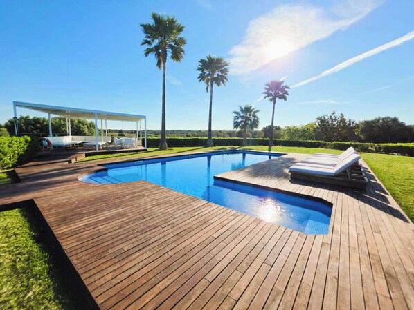 A swimming pool with a wooden deck, lounge chairs, and a covered seating area.