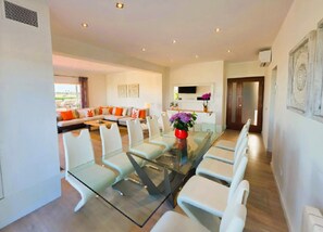 A modern living room with a glass dining table, white sofas, and a TV mounted on the wall.