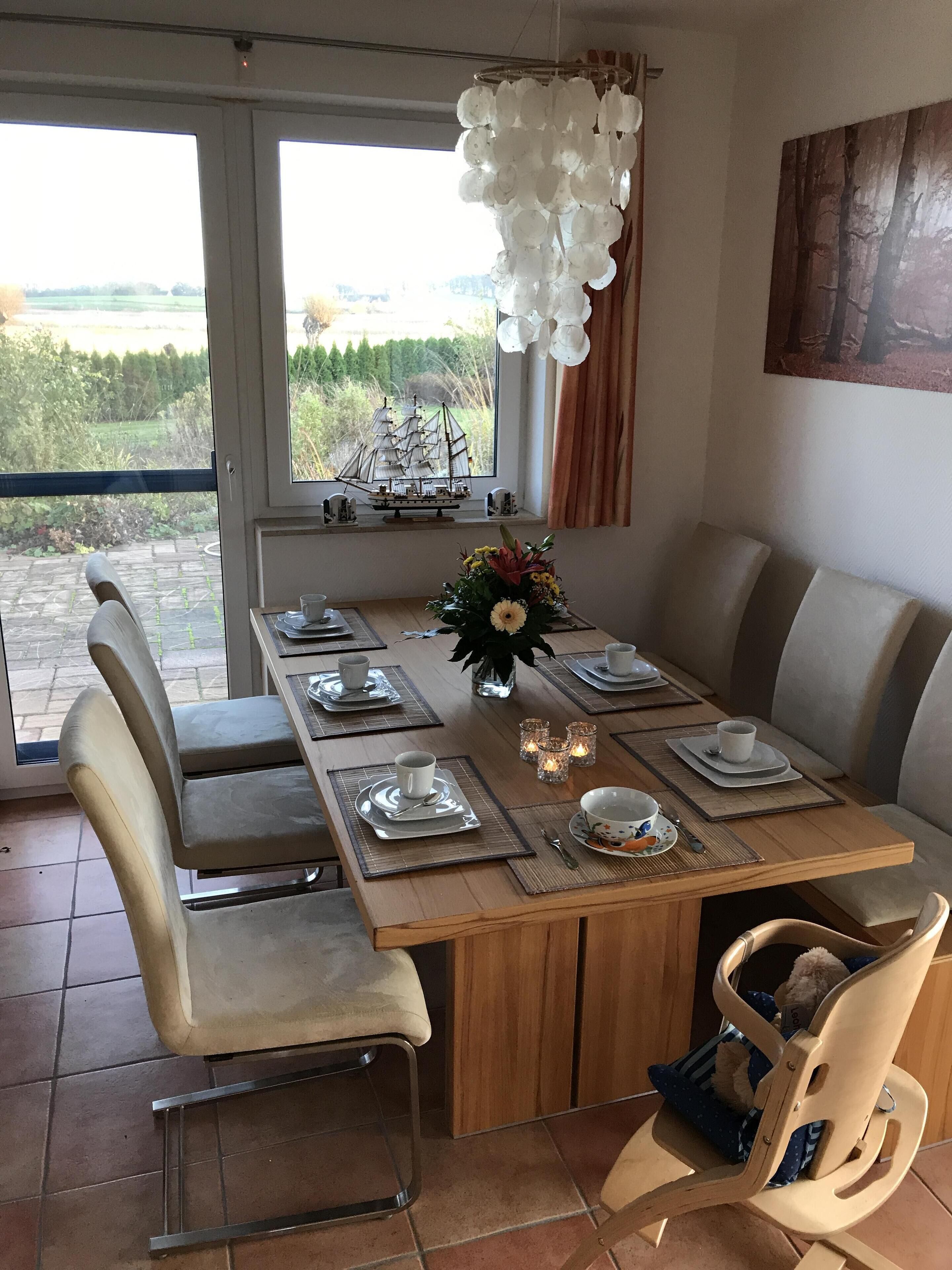 A dining area with a wooden table set for four, chairs, a vase with flowers, and a ship model on the windowsill.