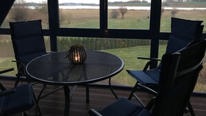 Ein überdachter Außenbereich mit einem runden Tisch und zwei Stühlen, eine Laterne auf dem Tisch sowie Blick auf einen See und Felder durch große Fenster.