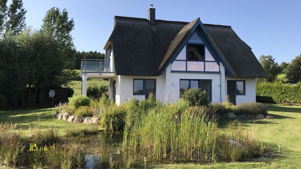 Ein Haus mit Strohdach, einem Balkon und einem Teich im Vorgarten.