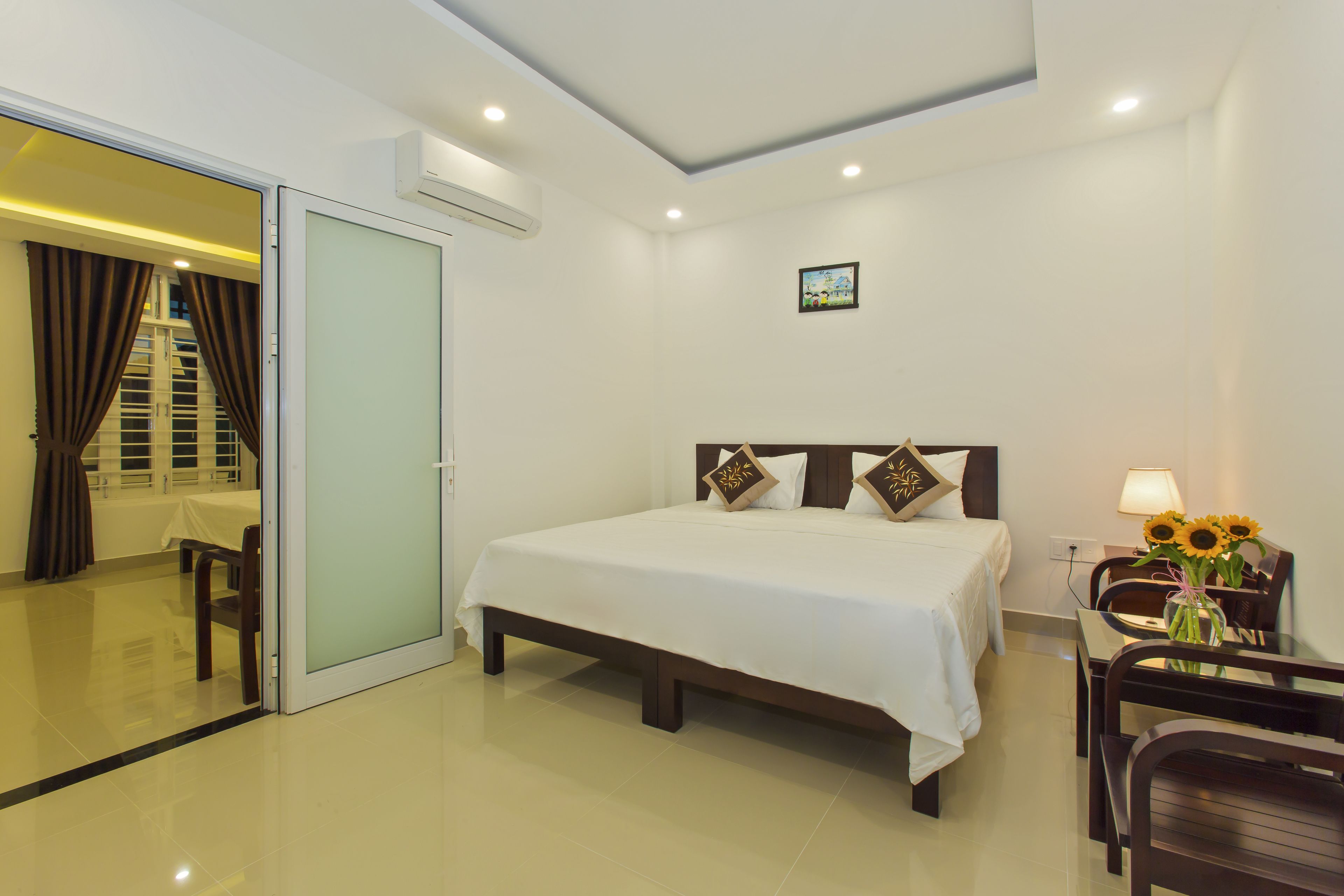 A hotel room with a bed, a desk, a chair, a vase with flowers, and a sliding door leading to a bathroom.