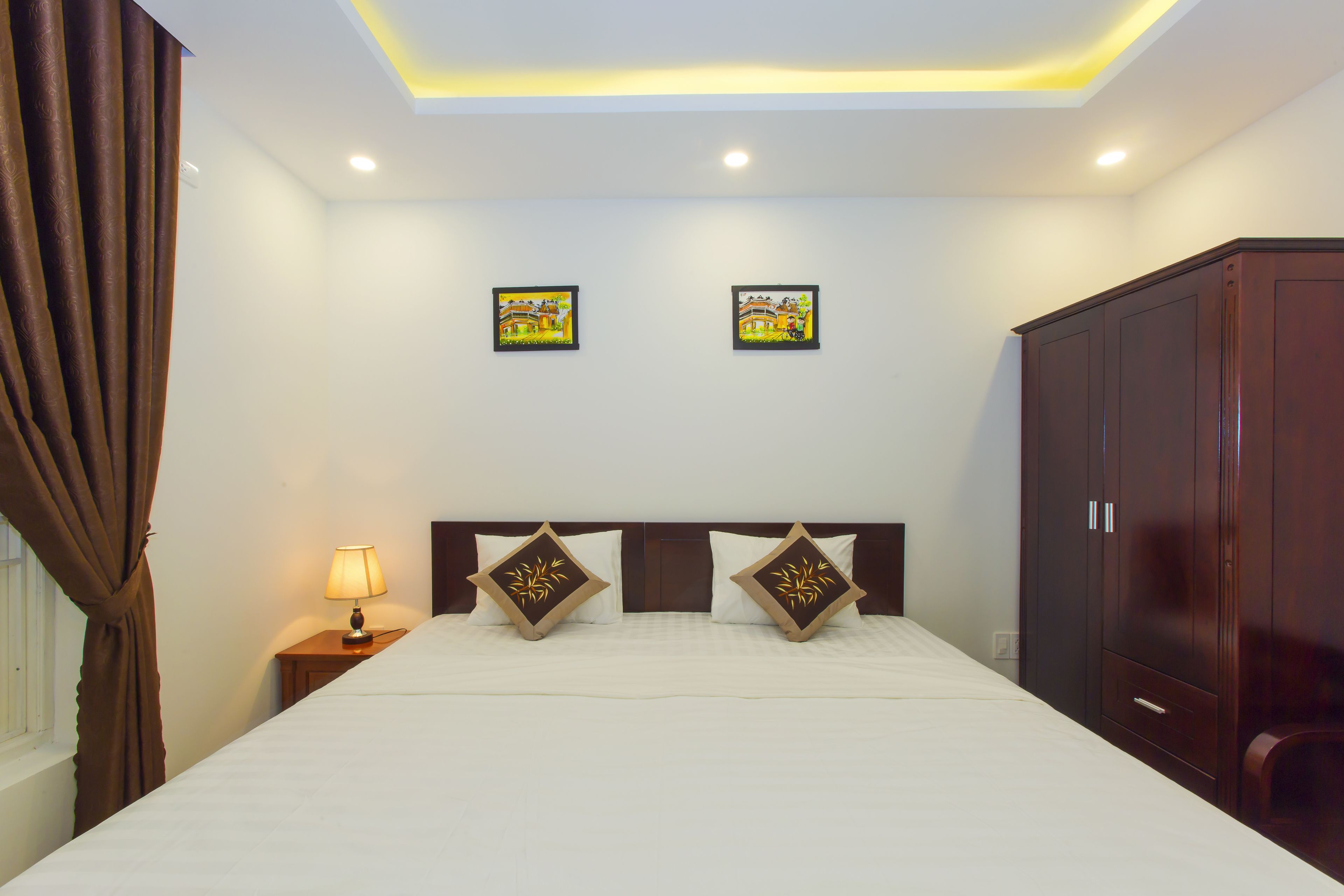 A hotel room with a large bed, two wall-mounted pictures, a nightstand with a lamp, and a wardrobe.