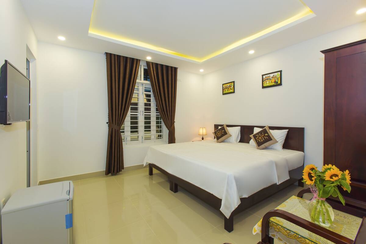 A hotel room with a bed, a TV, a window with curtains, a bedside table with a lamp, and a vase with flowers.