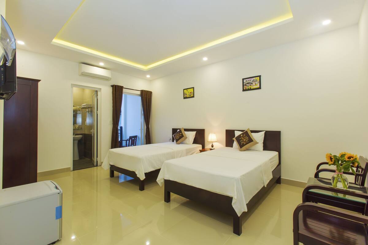 A hotel room with two beds, a desk with a chair, a TV, and a small table with a vase of flowers.