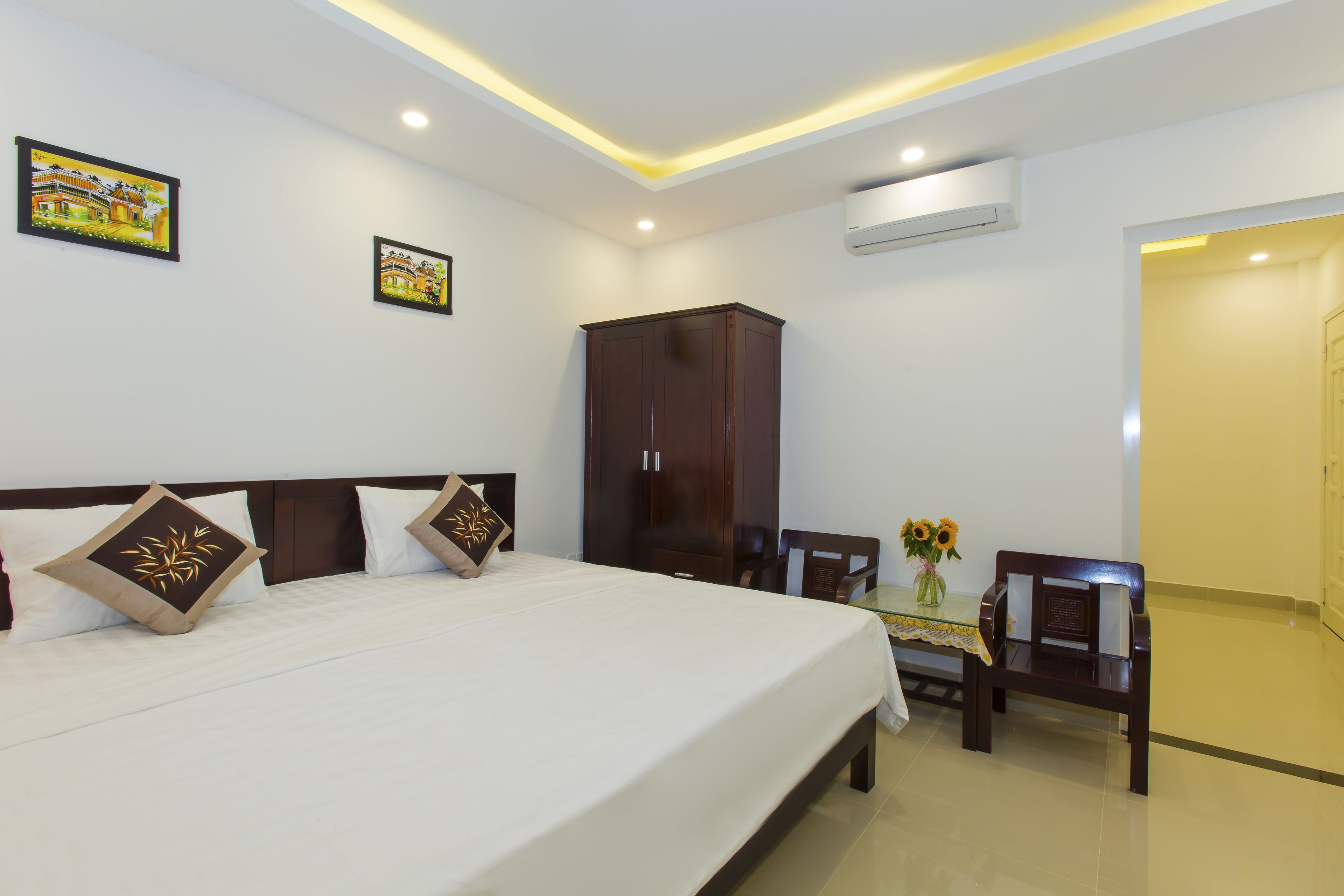 A hotel room with a large bed, a wooden wardrobe, a small table with a vase of flowers, and a bench.