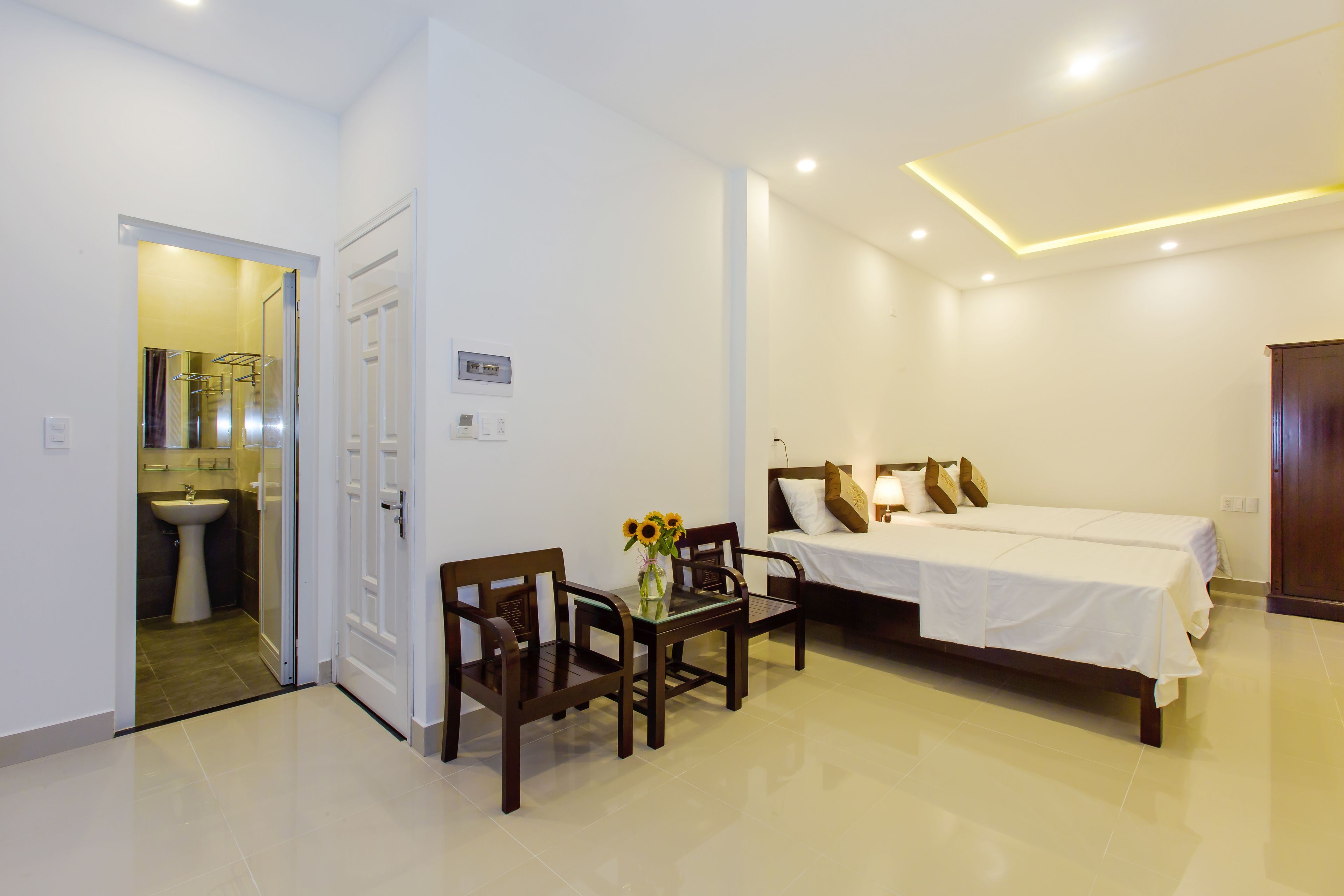 Une chambre d’hôtel avec un lit, une table et des chaises, un vase de fleurs et une salle de bain visible à travers une porte ouverte.