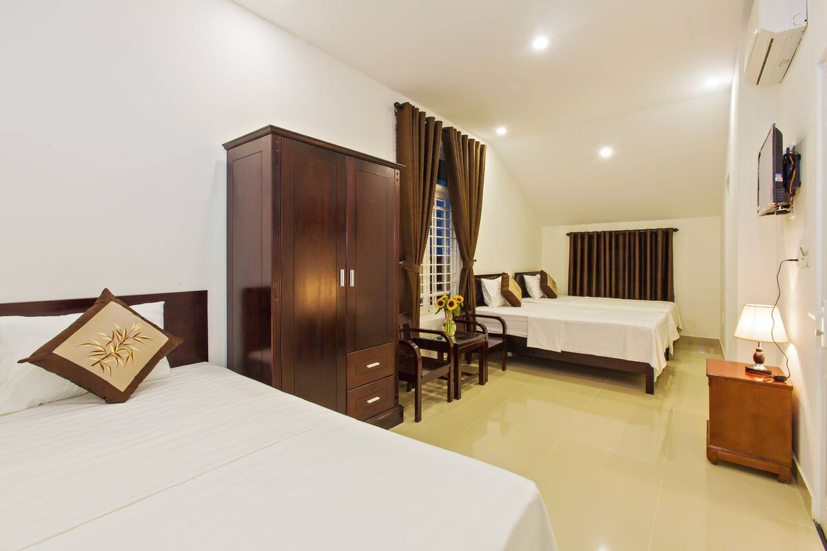 A hotel room with a large bed, a smaller bed, a wardrobe, a desk with a chair, and a nightstand with a lamp.