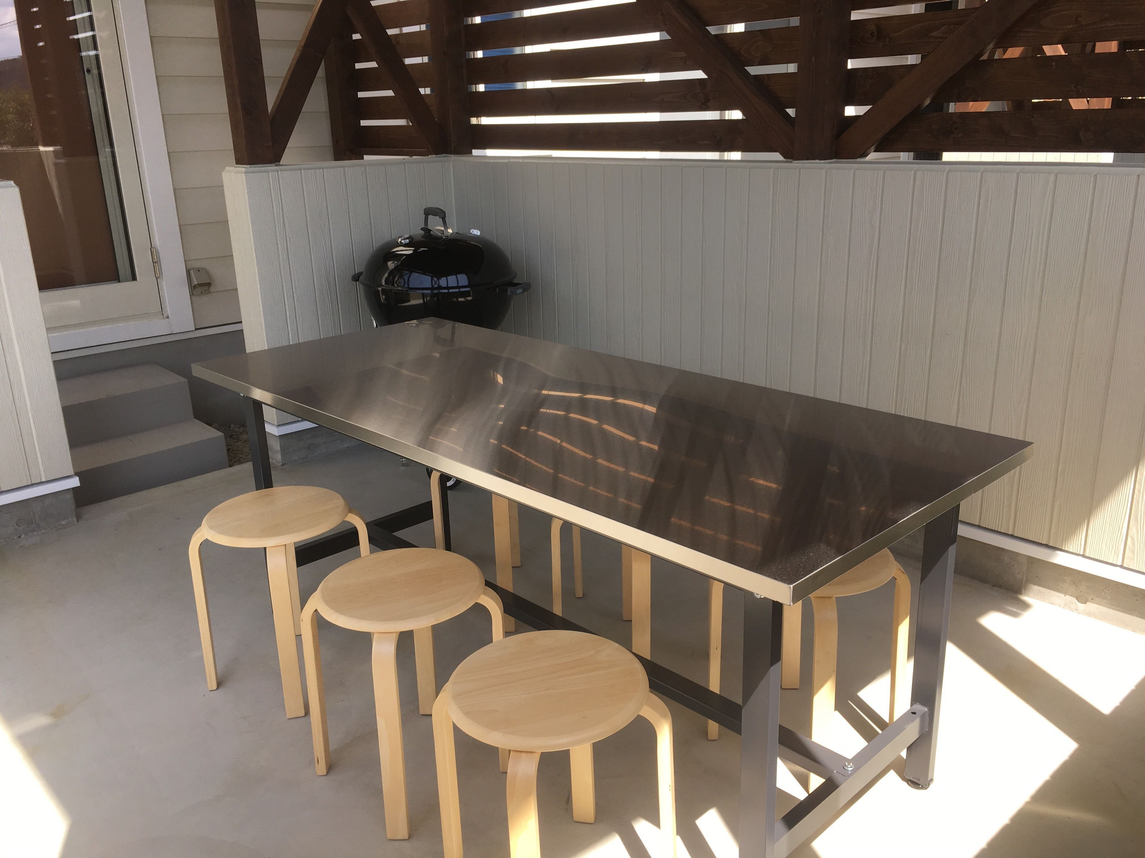 A modern outdoor dining area with a stainless steel table and four wooden stools.