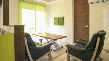 A room with two green chairs, a wooden table, a small rug, a window with green blinds, a door, and a wall-mounted TV.