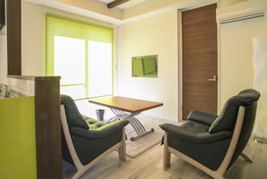 Quarto com duas cadeiras verdes, uma mesa de madeira, um tapete pequeno, uma janela com persianas verdes, uma porta e uma televisão fixada na parede.