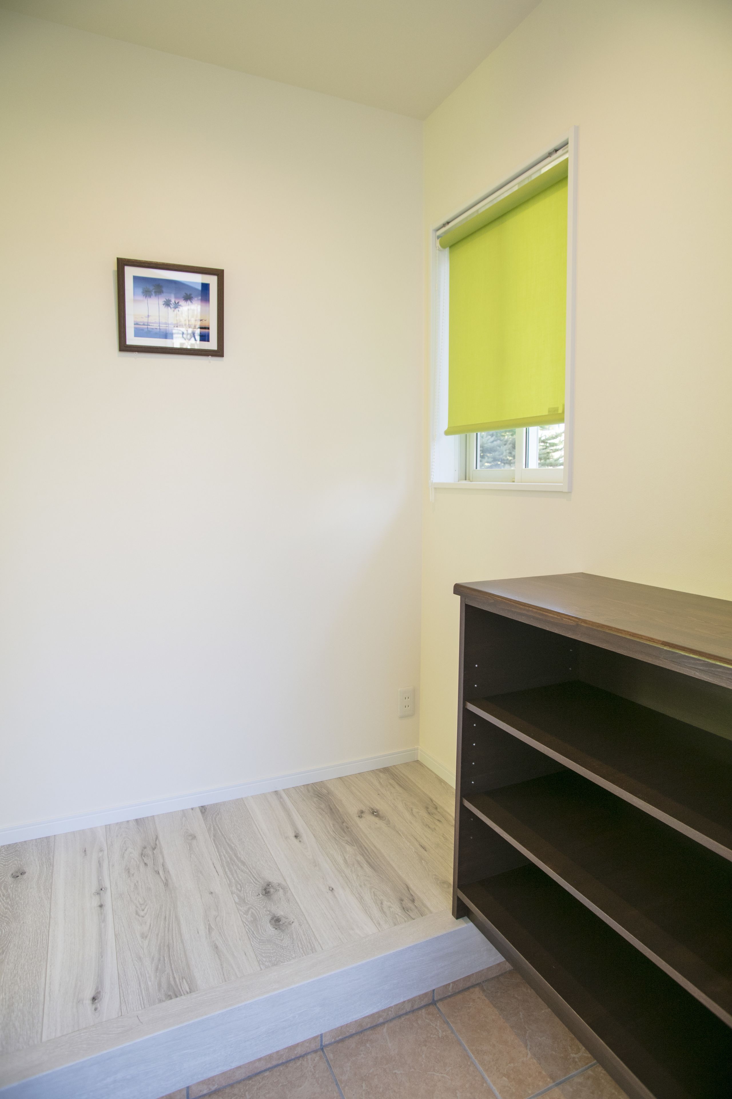 Een kamer met een lichte houten vloer, een raam met een geel zonnescherm, een lijst met een foto aan de muur, en een donker houten plankunit.