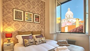 A neatly made bed with pillows, a bedside lamp, and a view of a building through a window. - Tejo I + II (Lisboa ciudad)