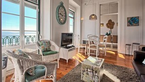 A spacious living room with a television, wicker chairs, a dining area, and a view of the sea. - Tejo I + II (Lisboa ciudad)