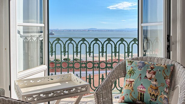 A balcony with a wicker chair and a small table, overlooking a coastal view with a metal railing and a distant cityscape. - Tejo I + II (Lisboa ciudad)