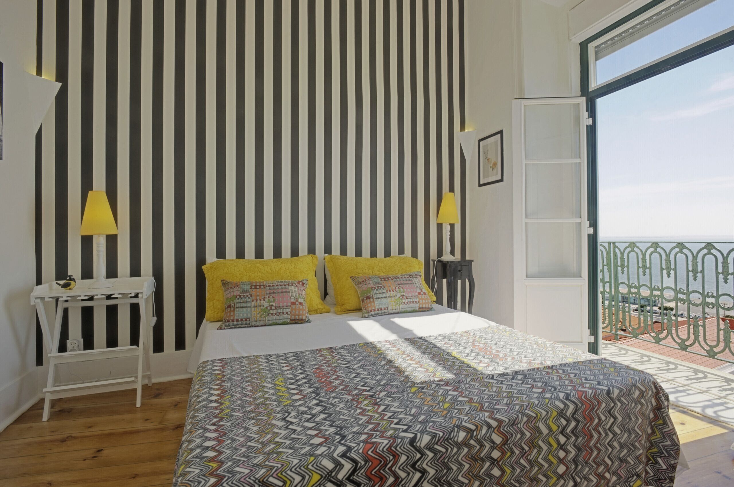 A bedroom with a striped wall, a bed with patterned bedding, two bedside lamps, and a view of a balcony with railings.