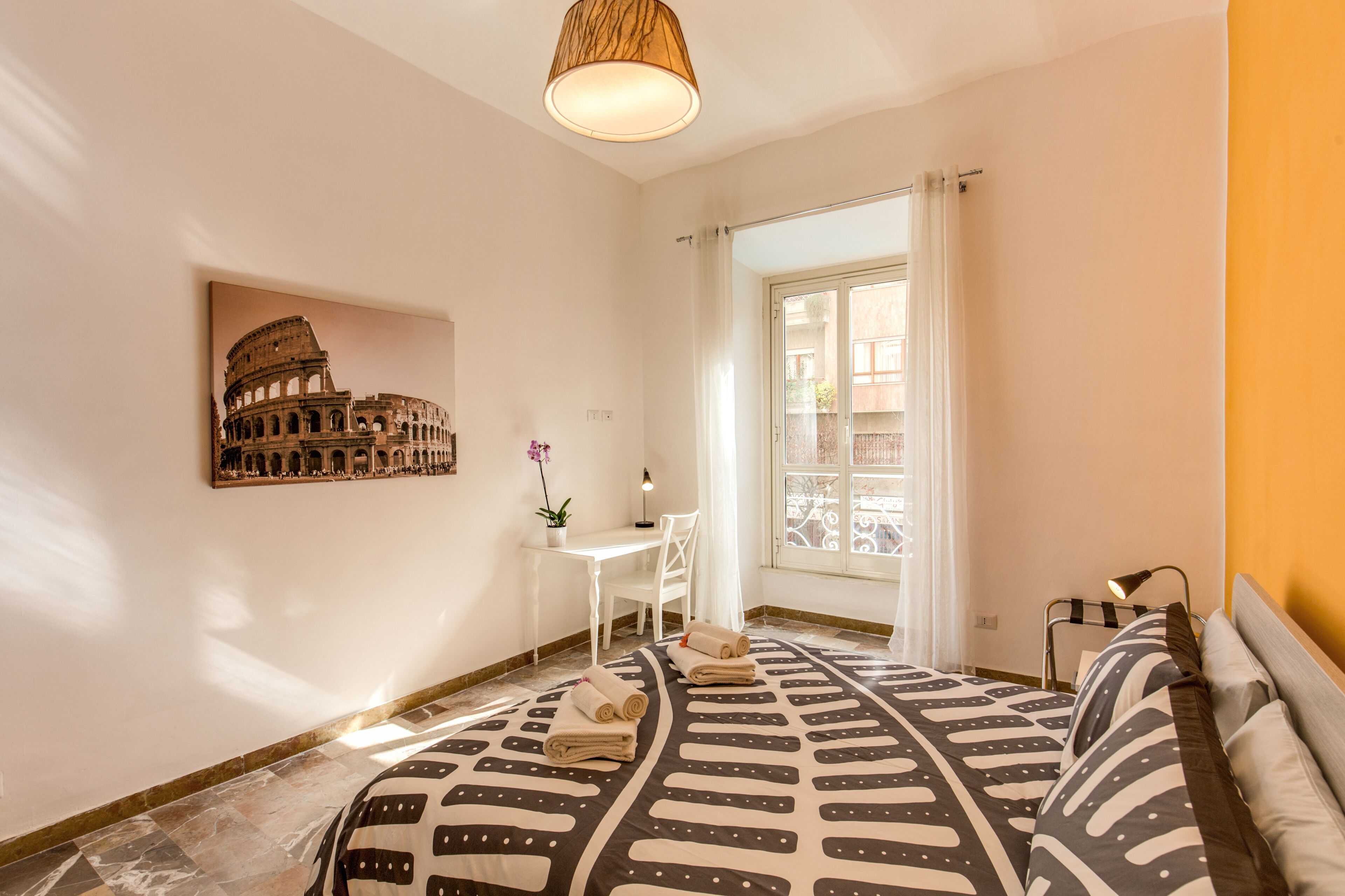 Ein Schlafzimmer mit Bett, Schreibtisch, Stuhl, Lampe und einem Fenster mit Vorhängen.