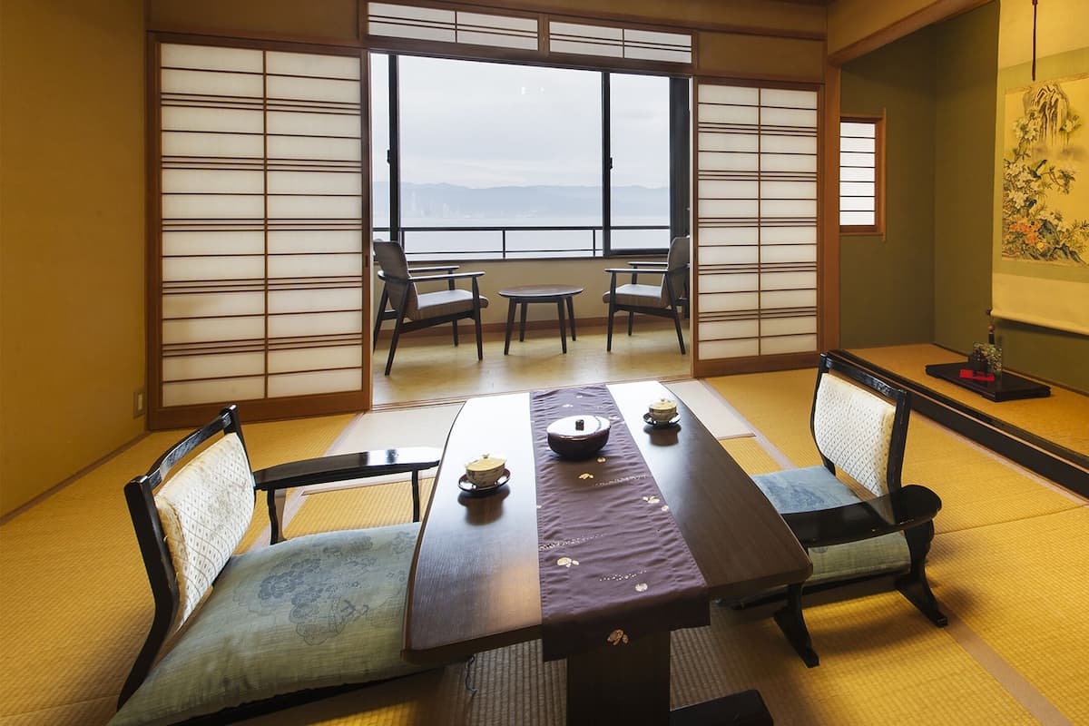 Una habitación japonesa tradicional con suelo de tatami, una mesa baja con cojines y vistas al exterior a través de biombos shoji.