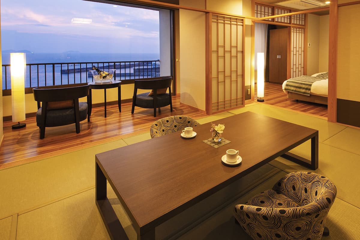 Una habitación tradicional japonesa con suelo de tatami, una mesa baja con sillas, una cama con futón y una puerta corredera que da a un balcón con vistas al mar.