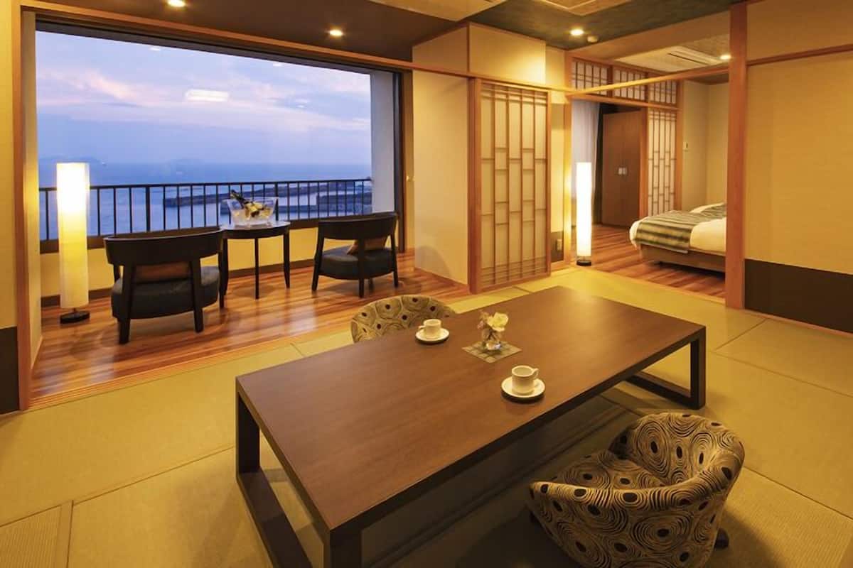 Una habitación japonesa tradicional con suelo de tatami, una mesa baja con sillas, una cama con futón y una puerta corredera.