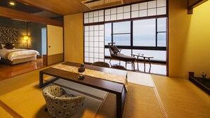 A traditional Japanese-style room with tatami flooring, a low table, a cushioned chair, and a bed with a wooden headboard. - Manpa Resort (Wakayama)