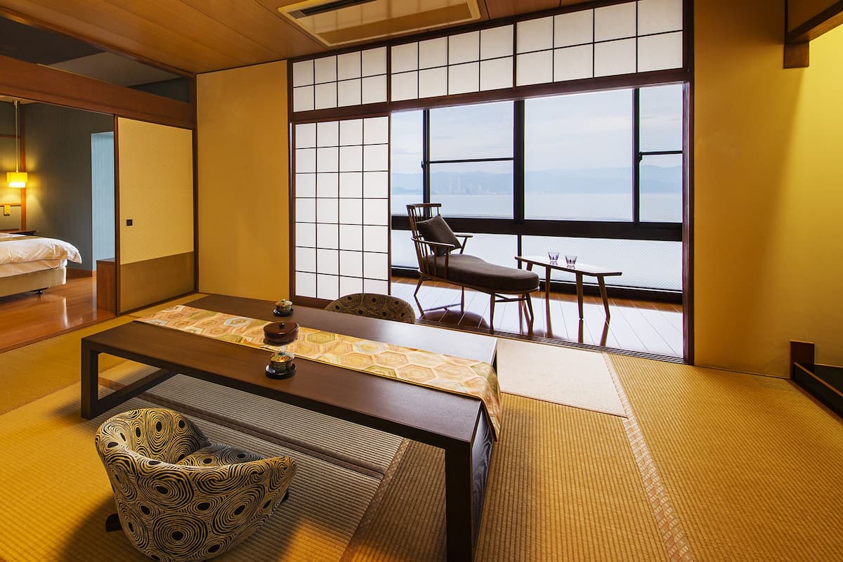 Una habitación de estilo japonés tradicional con suelo de tatami, una mesa baja, un sillón acolchado y una cama con cabecera de madera.