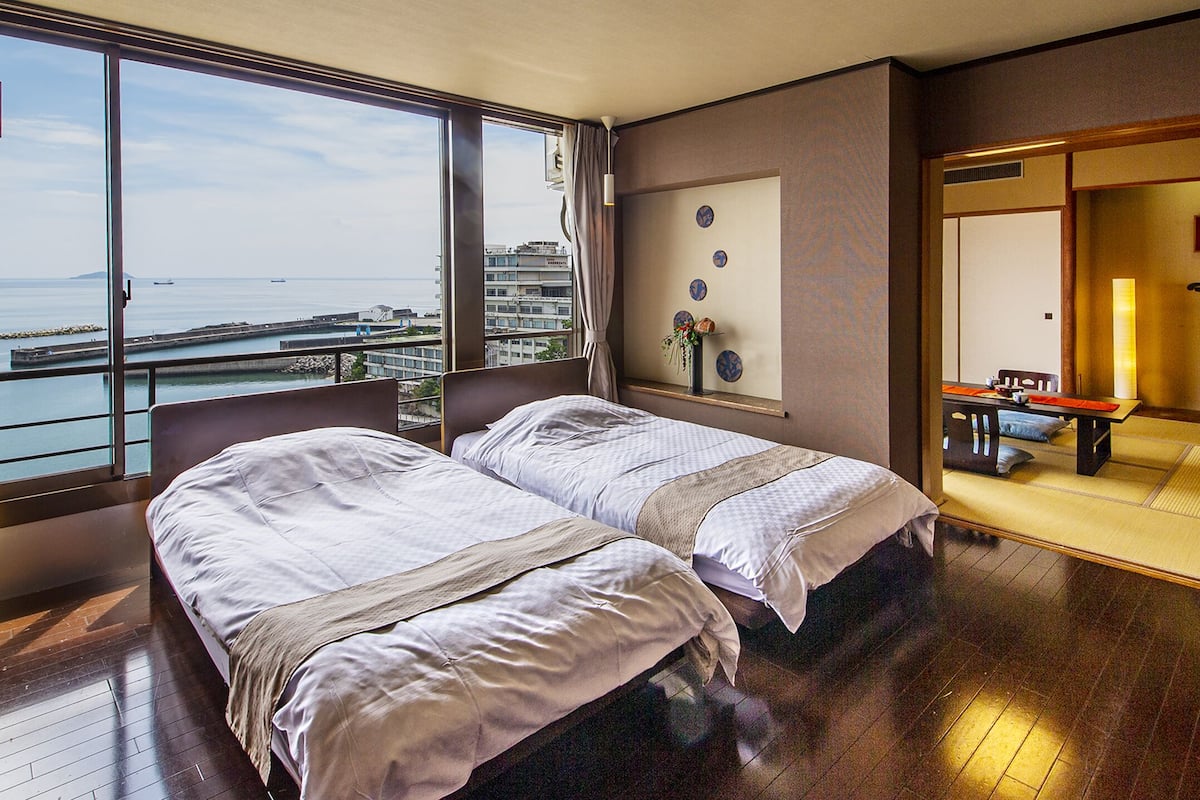 Habitación de hotel con dos camas, un ventanal con vista al mar, una mesita y un tradicional fusuma japonés.