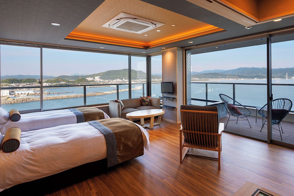 Una habitación de hotel moderna con dos camas, televisión, balcón con sillas y vista al mar.