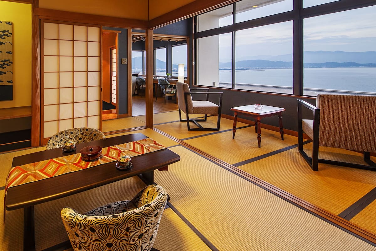 Una habitación tradicional japonesa con suelo de tatami, una mesa baja con un tapete estampado y vistas al mar a través de amplios ventanales.
