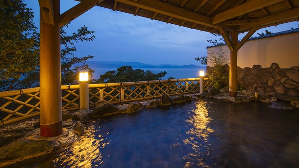 A tranquil outdoor hot spring area with a wooden deck, stone walls, and a view of the sea at dusk. - Manpa Resort (Wakayama)
