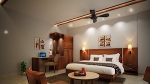 Una habitación de hotel moderna con una cama grande, un escritorio y una silla. Cuenta con un ventilador de techo, luces fijadas a la pared y cuadros enmarcados.