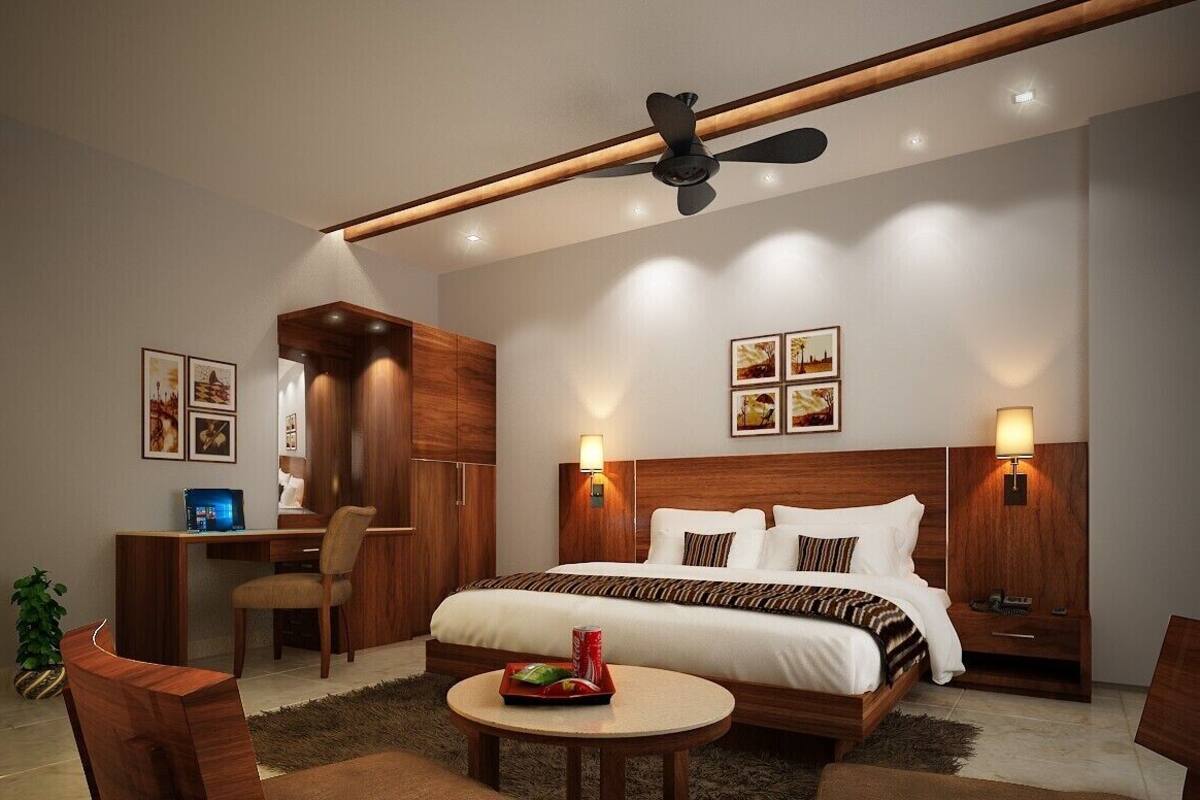 Ein modernes Hotelzimmer mit einem großen Bett, einem Schreibtisch und einem Sessel. Es gibt einen Deckenventilator, Wandleuchten und gerahmte Bilder an den Wänden.