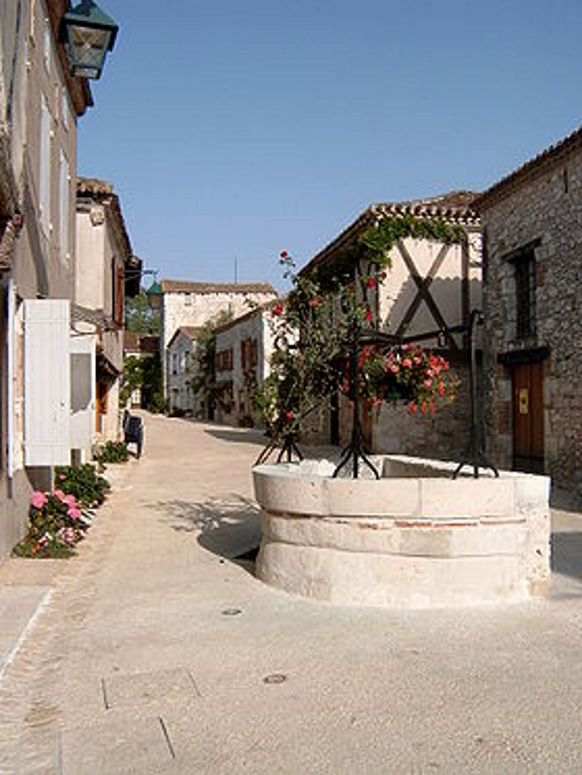 A narrow street lined with stone buildings, a central flower bed, and a person walking in the distance.