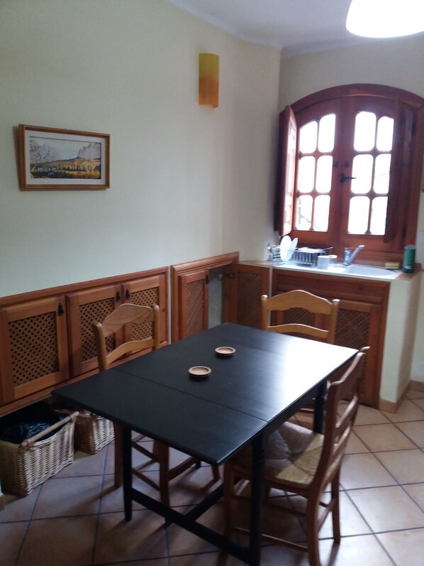 Un comedor con una mesa negra, sillas de madera, una cocina con ventana y un cuadro en la pared.