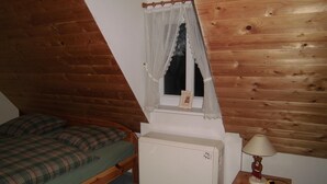 Une petite pièce chaleureuse avec un plafond en pente, un lit simple recouvert d’une couverture à carreaux, un radiateur blanc, une table de chevet avec une lampe et une fenêtre ornée d’un rideau en dentelle.