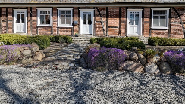 Une maison traditionnelle en briques, avec un chemin de pierre menant à l’entrée, entourée de gravier et de fleurs violettes.
